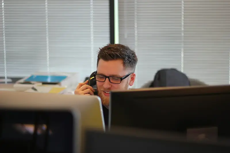 A smiling man in glasses talking on a desk phone handset in an office